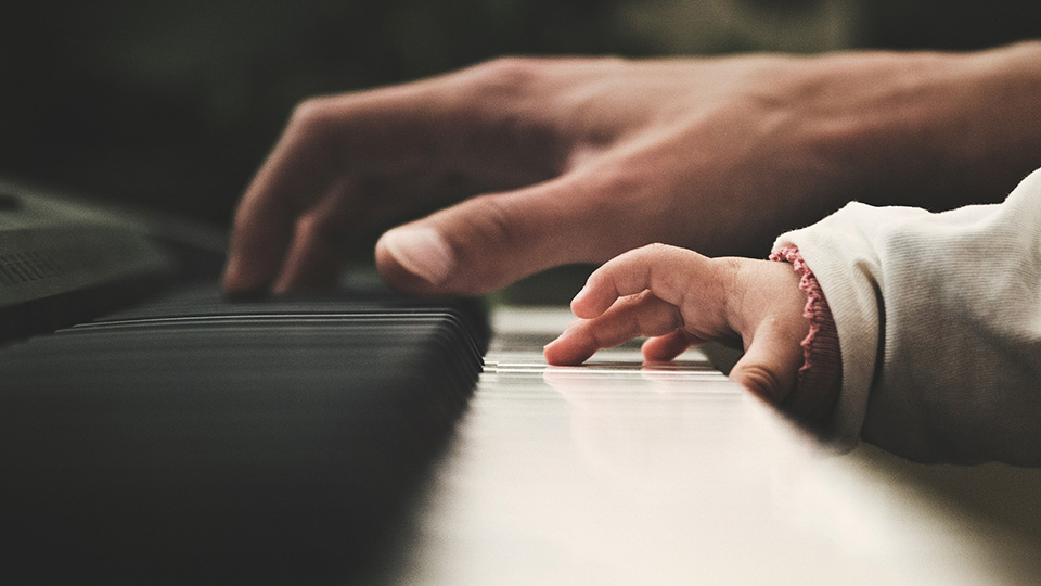 A close-up shot of a young child's hand, trying to play on a piano's white keys, with an adult hand in the background, instructing them.